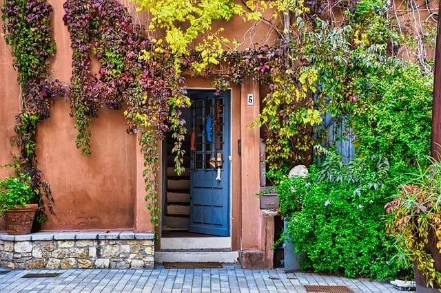 The image shows a rustic doorway with an open blue door, framed by vibrant autumn ivy in shades of red and green on an orange wall, conveying a warm, inviting mood.