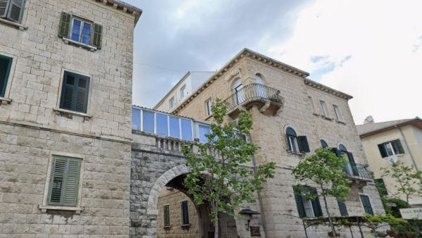 Accomodation Bacvice Split Affordable Split holiday rentalsy: A pale stone building in the Bačvice area features an arched stone doorway beneath a glassed-in walkway connecting two sections of the structure. Green-shuttered windows and a small wrought-iron balcony decorate the upper floors, with a young tree growing in front.