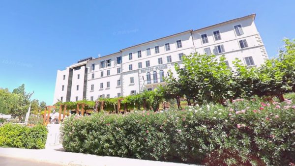 Bacvice, Split Affordable Split holiday rentals: A grand white hotel building with rows of shuttered windows stretches along a garden filled with blooming pink and white flowering shrubs and a leafy pergola near the Bačvice area. The scene is bathed in bright sunshine under a clear blue sky.