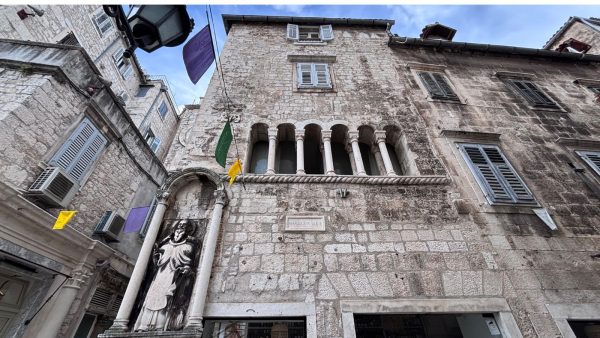 Beautiful Building in People's Square Split Affordable Split holiday rentals: A tall stone building on People's Square rises above a row of slender columns forming an open gallery with arched windows on the upper level. In the foreground, a carved statue of a robed figure stands on a stone pedestal beside the entrance, with purple and yellow flags fluttering overhead.