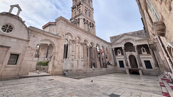 Diocletian's Palace Split, Croatia Affordable Split holiday rentals: Inside the ancient Peristyle courtyard of Diocletian's Palace, rows of tall stone columns with decorative capitals line an open plaza beneath a towering bell tower. The worn stone pavement stretches toward a set of steps, with a lone pigeon resting on the ground and red cushioned seats visible to the right.