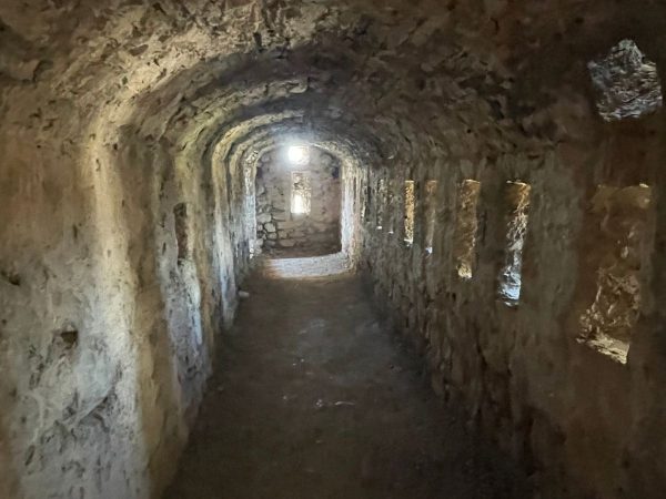 Inside the Klis Fortress Walls Split Day trips from Split. A dim stone corridor inside Klis Fortress, with an arched ceiling and narrow rectangular openings cut into the thick walls to let in slivers of light. The passage curves toward a small sunlit window at the far end, with the stone floor and walls showing centuries of wear.
