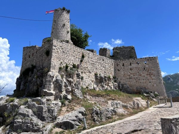 Klis Fortress Split Croatia Day trips from Split. A view from below of Klis Fortress perched on a rocky outcrop, its pale stone walls topped with a round tower flying the red and white checkered Croatian flag. A small tree grows within the fortress walls, with a paved path leading up to the entrance under a bright blue sky with scattered clouds.