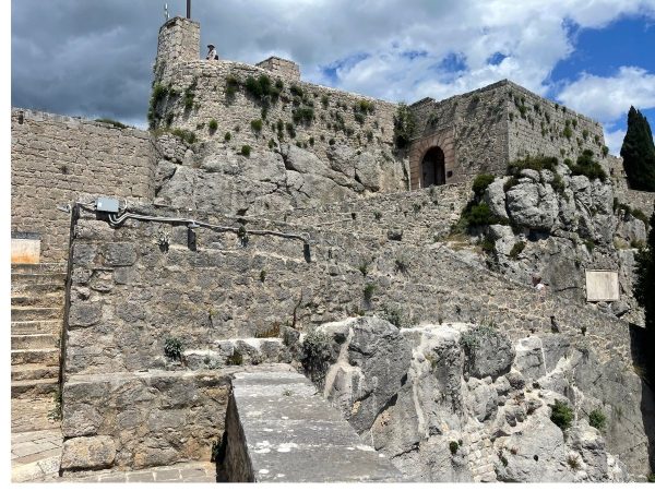 Klis Medieval Fortress Split Croatia Day trips from Split. A close-up view of Klis Fortress's weathered stone ramparts, with wild plants growing between the rocks and a person standing at the top near a tower. A stone archway entrance is visible on the right, with a cypress tree nearby and dramatic storm clouds gathering in the sky above."