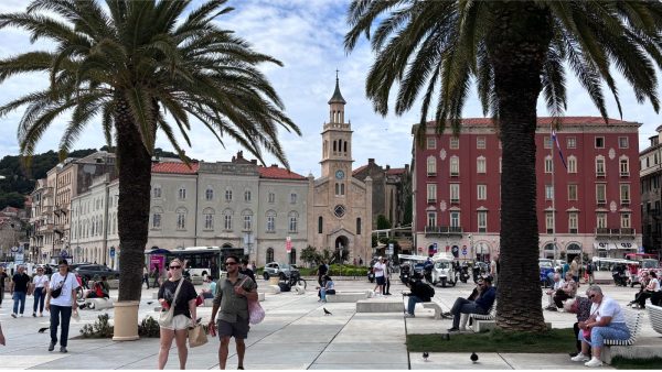Looking Toward the Veli Varoš District in Split Affordable Split holiday rentalsy: Two tall palm trees frame a view of a historic square featuring a slim clock tower flanked by a cream-colored building and a deep red building with white-trimmed windows. Pedestrians walk and sit on benches in the foreground, with pigeons scattered across the paved plaza.