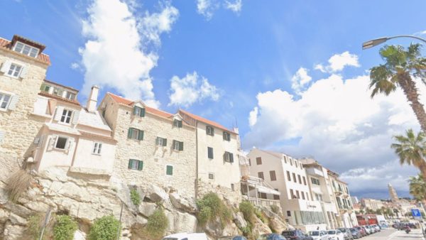 Meje Apartment, Split Affordable Split holiday rentals: A cluster of pale stone houses with green-shuttered windows and red tile roofs perches on a rocky cliff edge in the Meje district. A row of parked cars lines the road below, with a palm tree and glimpses of the city and a distant white tower visible under a bright blue sky with fluffy clouds.