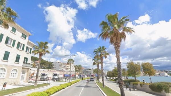 Meje, Split Affordable Split holiday rentals: A wide palm-lined boulevard curves along the coast in the Meje neighborhood, with a stone building on the left and the turquoise sea visible on the right. Construction trucks are parked along the road, and a few pedestrians walk beneath the palm trees toward the city skyline in the distance.