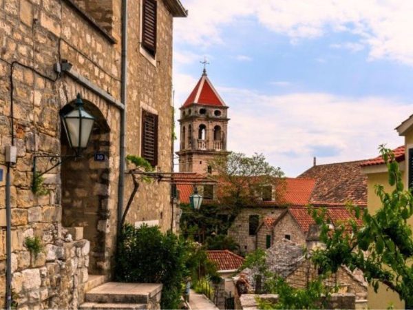 Omis Old Town Day trips from Split. A narrow stone street in Omiš's Old Town, lined with weathered stone buildings and a hanging lantern. A church bell tower with a red conical roof rises above terracotta-roofed houses in the background, with green vines and plants growing along the street under a partly cloudy sky.