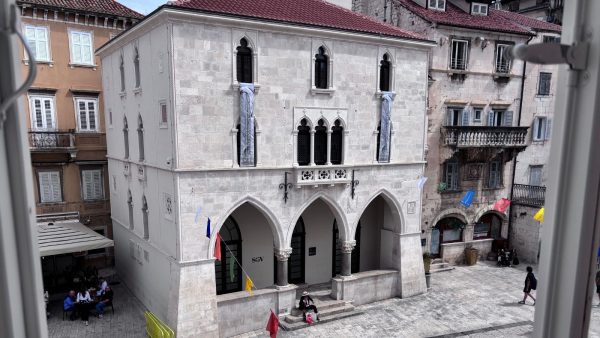 _People's Square, known locally as Narodni trg or simply Pjaca. Affordable Split holiday rentals: Viewed through an open window, this photo shows a Gothic-style stone building with pointed arched windows and a triple-arched arcade at street level on People's Square, also known locally as Narodni trg or Pjaca. Below, people sit at outdoor café tables and walk across the square's smooth stone pavement.