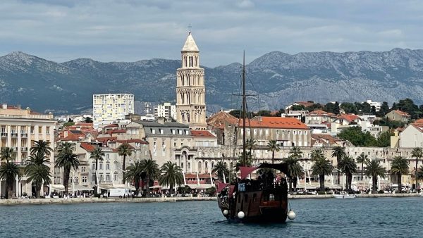 Split Croatia From the Water Affordable Split holiday rentals: A view of the Split waterfront seen from the sea, with a tall stone bell tower rising above a row of palm trees and pastel historic buildings with terracotta roofs. In the foreground, a wooden sailing boat with dark red sails and white fenders drifts across the calm blue water, with rugged gray mountains in the background.