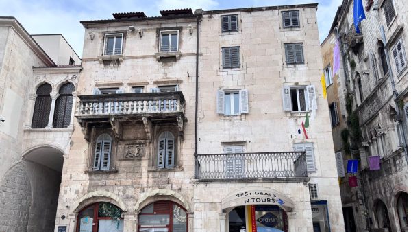 Stunning Apartments in People's Square Split Affordable Split holiday rentalsy: A weathered stone apartment building on People's Square features an ornate stone balcony with carved supports and shuttered windows on the upper floors. At street level, shopfronts including a travel agency and a bistro sit beneath the historic facade, with colorful flags hanging nearby.