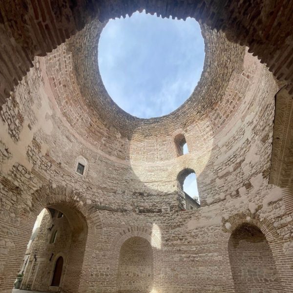 Split Best Places to Stay: Looking straight up inside the ancient domed Vestibule of Diocletian's Palace, a large circular opening at the top reveals a patch of blue sky with wispy clouds. The weathered stone and brick walls curve upward in rings, with several small arched windows and doorways visible around the base of the chamber.