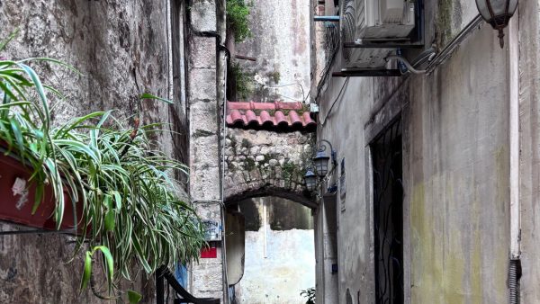 Meandering Through the Back Streets of Split Affordable Split holiday rentals. A narrow passageway of old stone buildings with a row of green plants on the left. At the end of the alley up high is enclosed walkway between the two buildings.