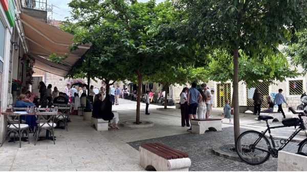 Vibrant Veli Varoš District Affordable Split holiday rentals: A shaded pedestrian square in the lively Veli Varoš district is filled with leafy green trees, stone benches, and people gathered outside cafés under a tan awning. A bicycle leans against one of the tree planters while a small child runs across the paved walkway.