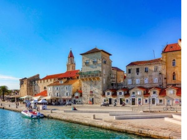 Trogir Promenade Croatia Day trips from Split. A sun-drenched waterfront promenade in Trogir, Croatia, lined with historic stone buildings, red-tiled roofs, and a pointed cathedral spire. Small boats moor along the paved quay beside sparkling turquoise sea water, with palm trees and outdoor cafés stretching beneath a bright blue sky.