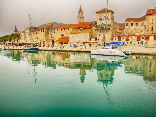 Trogir Stunning Waterfront Day Trips from Split. A tranquil, golden-toned view across clear emerald waters reflecting the historic waterfront of Trogir, Croatia. White catamarans and boats float near the stone promenade, where red-tiled roofs, medieval towers, and a pointed cathedral spire rise under a soft, overcast sky.
